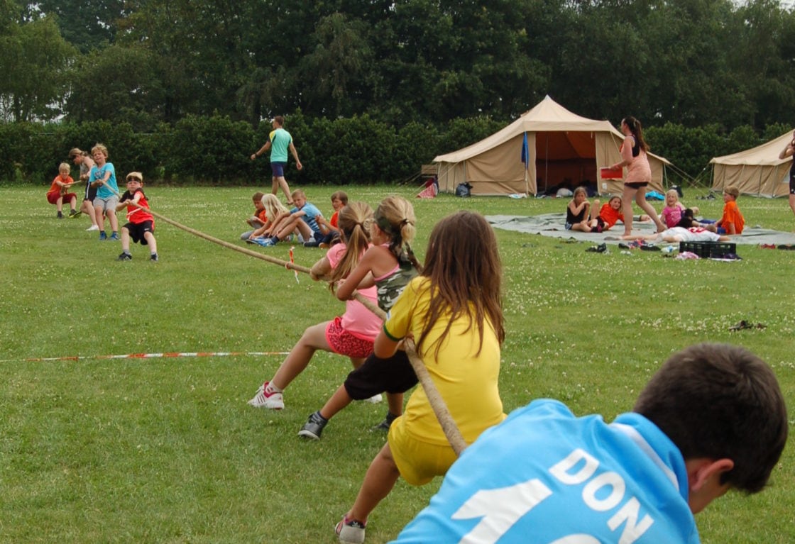 Kinderen doen touwtrekken op een zomerkamp van Jong Nederland met tenten op de achtergrond