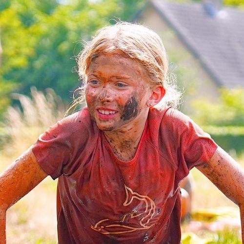 Een lachend meisje onder de modder tijdens een spel in de buitenlucht bij Jong Nederland