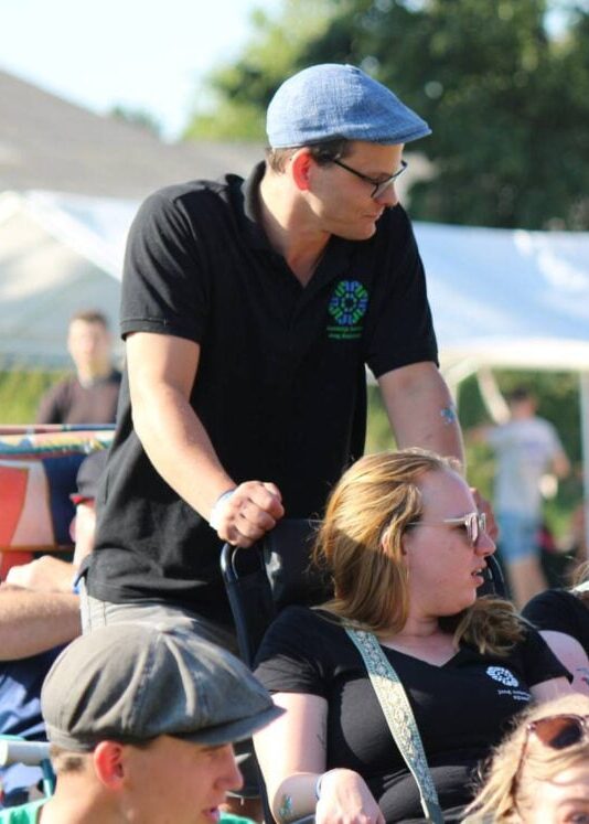 Vrijwilliger in poloshirt van Jong Nederland duwt een rolstoel over het zonnige festivalterrein